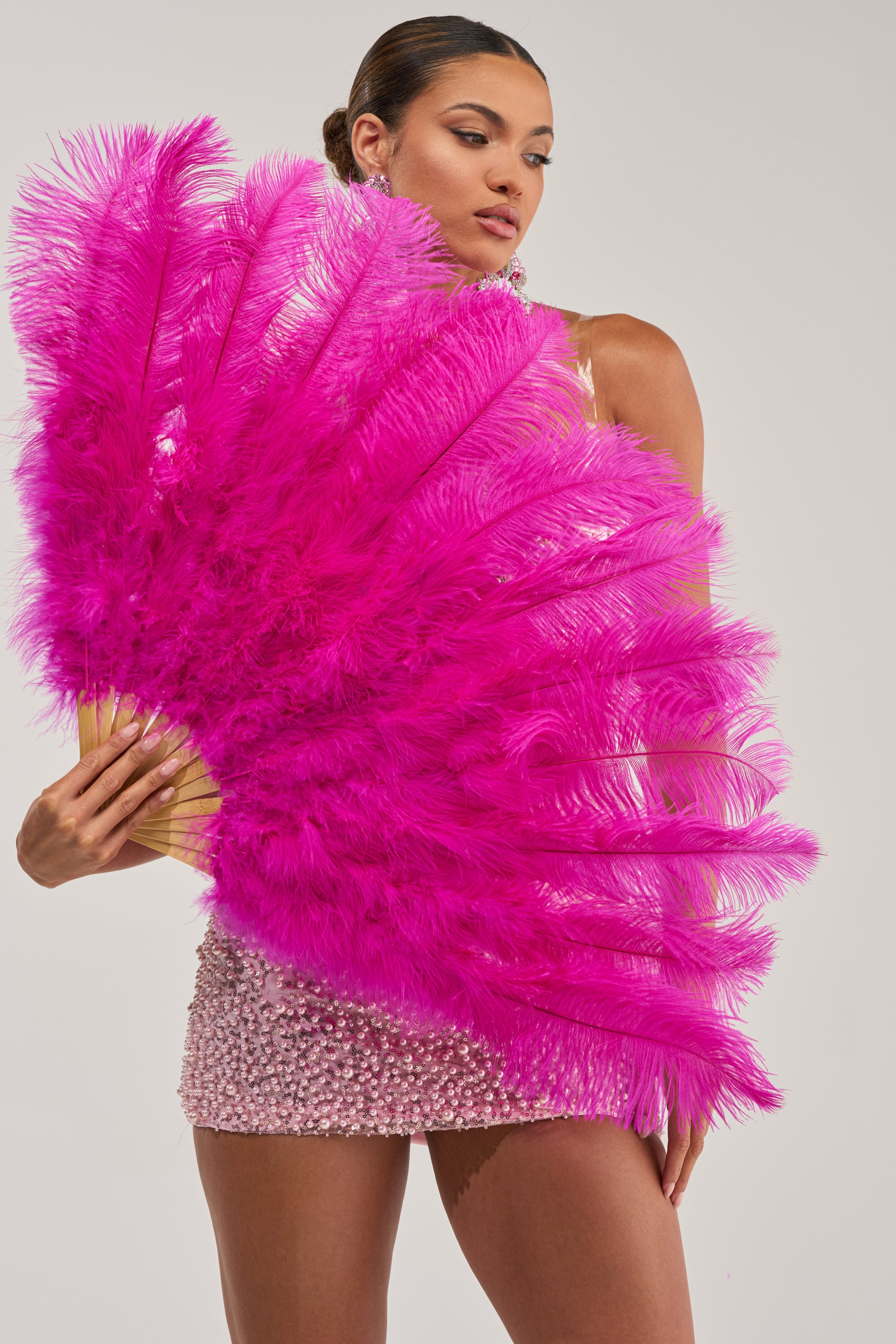 A woman in a beaded mini dress confidently poses with the STAY THE NIGHT FEATHER FAN, its hot pink ostrich feathers partially covering her against a light background—a bold choice for standing out at any party.