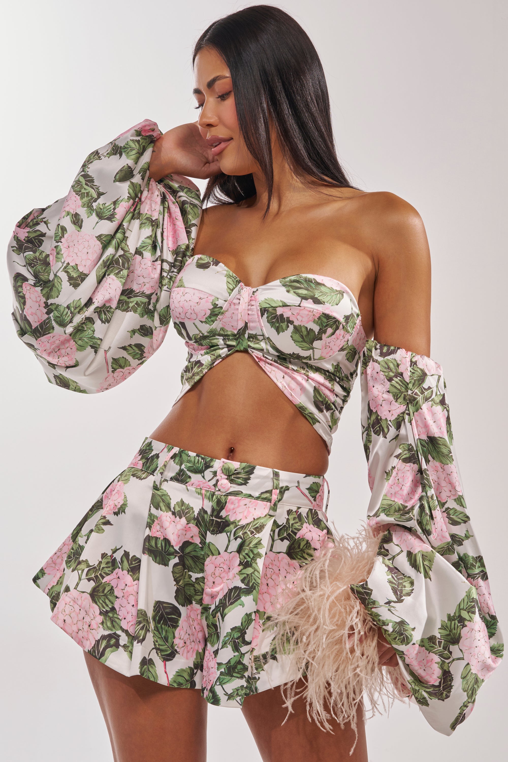A woman smiles as she poses in the ONE OF A KIND FLORAL BLOUSE, featuring pink flowers and green leaves, styled with a short skirt. The blouse has puffy off-the-shoulder sleeves, and she holds a small feathered bag. 1