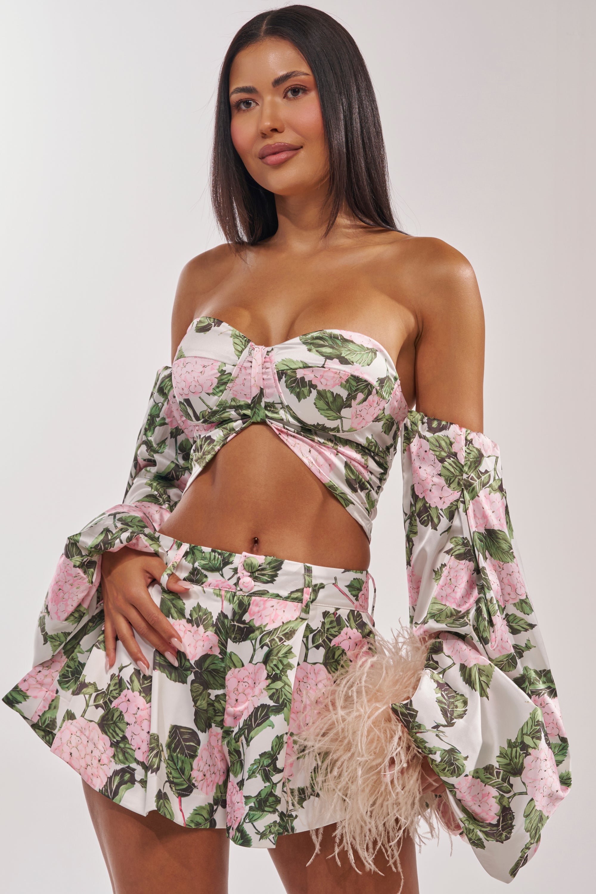 A woman wears the ONE OF A KIND FLORAL BLOUSE two-piece set with pink and green floral patterns, a strapless crop top with flared sleeves, matching high-waisted shorts, and holds a beige feathered accessory. 2