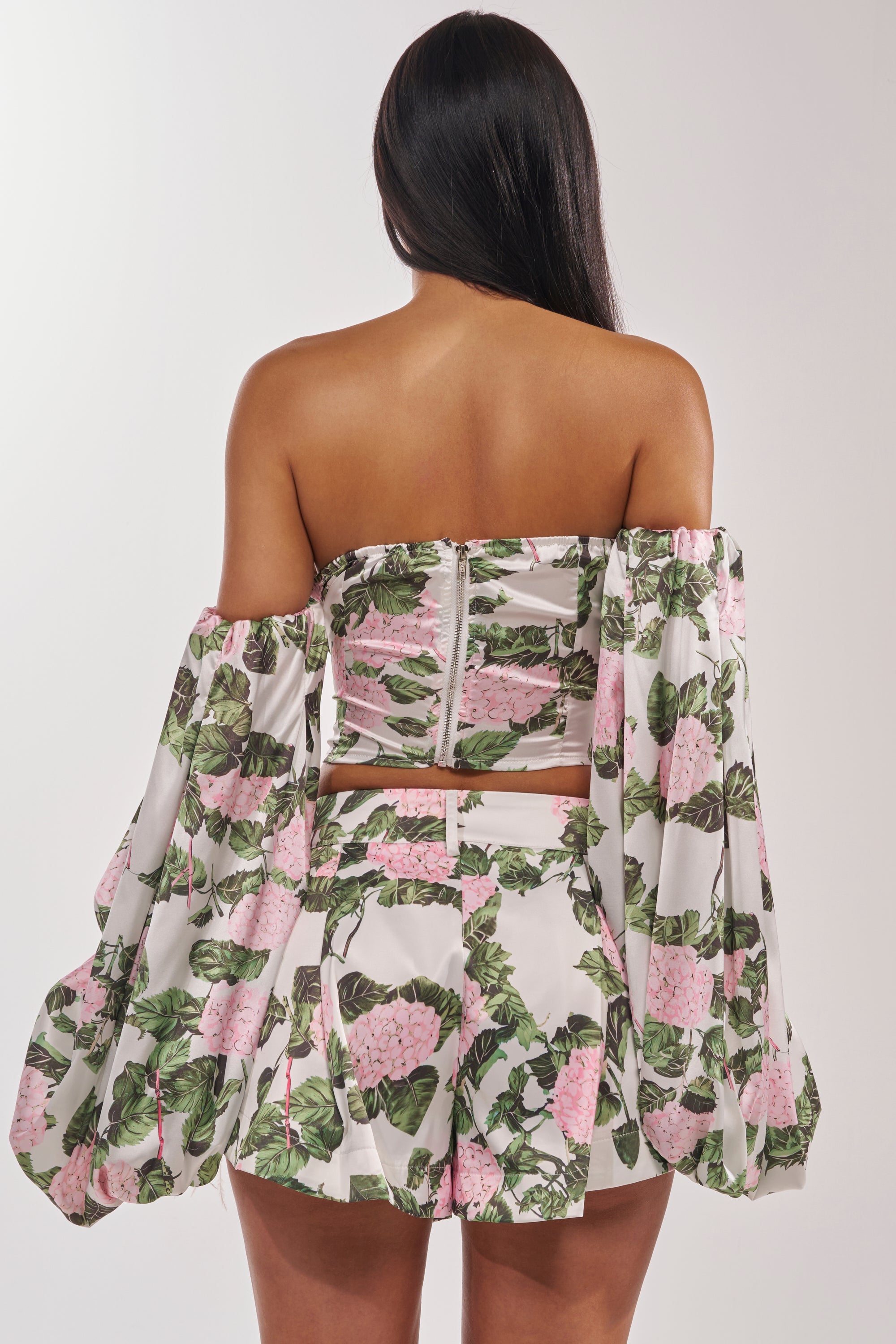 A woman with long dark hair is seen from the back wearing the ONE OF A KIND FLORAL BLOUSE with puffed off-the-shoulder sleeves and pink flower and green leaf prints on a white background, paired with matching high-waisted shorts. 4