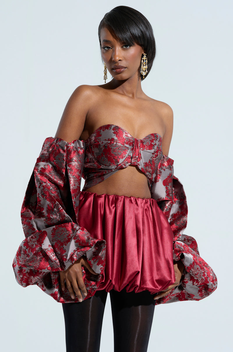 A woman poses confidently, wearing the DAISY OFF THE SHOULDER BLOUSE—a stylish red piece with voluminous sleeves and a cropped silhouette, beautifully adorned with a floral metallic pattern. Her hair is sleek, and she accessorizes with statement earrings against a plain light gray background.