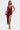A woman wears the SOFIA FRINGE HIGH SLIT MIDI DRESS IN BURGUNDY, featuring a sleeveless design, draped neckline, and high slit. She holds a gold clutch and wears strappy gold heels against a plain white background.