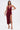 A woman stands against a white background wearing the SOFIA FRINGE HIGH SLIT MIDI DRESS IN BURGUNDY, featuring a sleeveless design, cowl neckline, and high slit. She holds a gold clutch and wears gold high-heeled sandals.