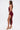 A woman stands in profile against a plain white background wearing the SOFIA FRINGE HIGH SLIT MIDI DRESS IN BURGUNDY, a sleeveless, textured dress, paired with gold strappy sandals.