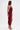 A woman with straight brown hair in gold high-heeled sandals poses sideways in the SOFIA FRINGE HIGH SLIT MIDI DRESS IN BURGUNDY, featuring a sleeveless design, deep neckline, and high side hem, set against a plain white background.