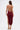 A woman with long brown hair wears the SOFIA FRINGE HIGH SLIT MIDI DRESS IN BURGUNDY, paired with gold high-heeled sandals and a gold clutch, standing against a plain white background.