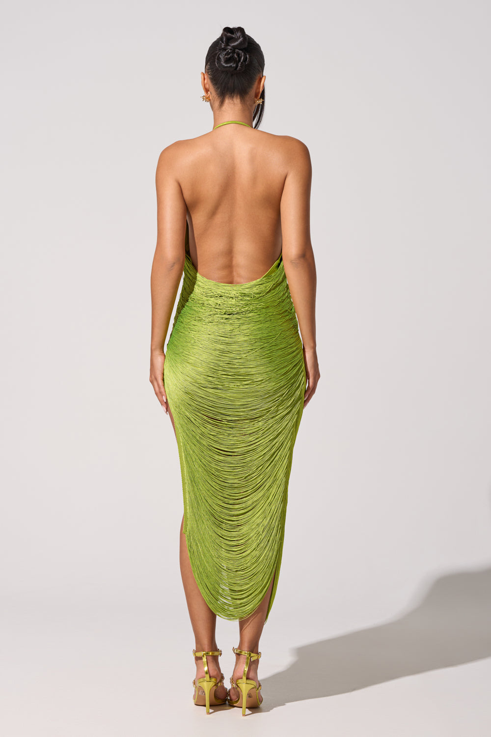 A person faces away, wearing the SOFIA FRINGE HIGH SLIT MIDI DRESS IN GREEN with a neat bun and strappy yellow heels against a plain light background—a chic, standout look for parties. 6