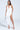 A woman models the GIANINA FRINGED COWL NECK MAXI DRESS IN WHITE. Sleeveless with an open back and cascading fringe tassels, she pairs it with a silver handbag, silver high heels, flowing long hair, and hoop earrings against a plain background.