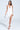 Someone confidently walks in a flowing AKIRA Label GIANINA FRINGED COWL NECK MAXI DRESS IN WHITE featuring a high slit. They carry a small metallic handbag and wear silver high-heeled sandals against a plain, light background.