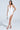 A person in a white AKIRA Label Gianina Fringed Cowl Neck Maxi Dress with a deep neckline and high slit, holding a small silver purse. They have long hair, wearing silver high-heeled sandals against a plain light-colored background.