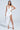 A woman poses confidently in a GIANINA Fringed Cowl Neck Maxi Dress in white, featuring a deep plunge and high slit. She holds a silver clutch and wears silver heels. Her straight dark hair contrasts the plain gray background.
