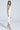 Wearing the GIANINA Fringed Cowl Neck Maxi Dress in White, a woman stands gracefully against a plain background. Her silver high heels sparkle as she holds a small purse and gazes at the camera, with long brown hair cascading over her shoulders.