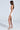 A woman poses in profile in the GIANINA Fringed Cowl Neck Maxi Dress in White, featuring a high side slit. Her long brown hair enhances her look as she pairs it with silver high-heeled sandals against a plain, well-lit background.