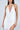 A person in a sleeveless Gianina Fringed Cowl Neck Maxi Dress in white with thin straps, deep cowl neckline, and high slit holds a metallic clutch. Their hair is down with hoop earrings against a plain gray background.