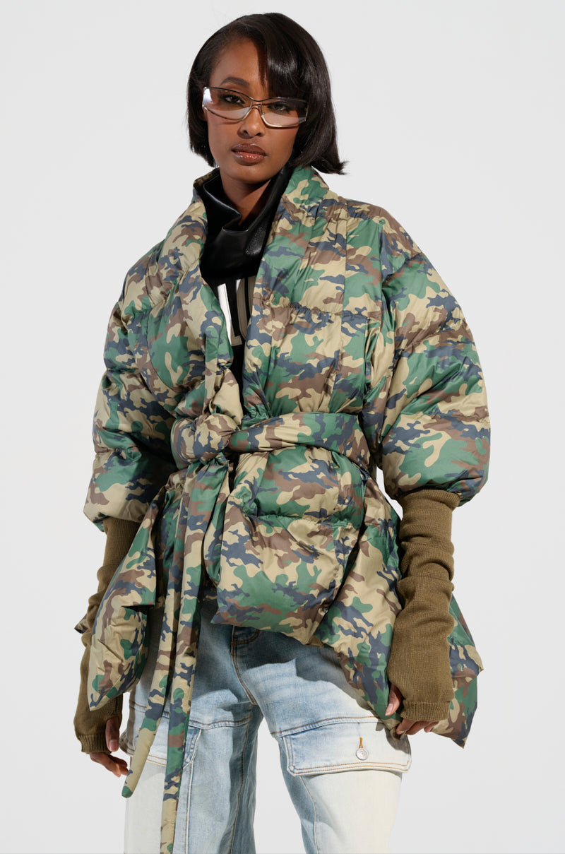 A person wears the Camo Long Rib Sleeve Peplum Puffer Coat, which features an oversized fit and a belt. They pair it with light blue jeans and long brown gloves, adding sunglasses and short, stylish hair to complete the look.