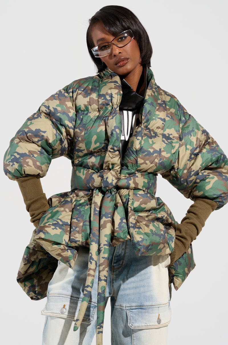 A person confidently poses in the CAMO LONG RIB SLEEVE PEPLUM PUFFER COAT and blue jeans. They accessorize with rectangular glasses, placing their hands on their hips to highlight the cinched waist design and ribbed sleeves of the jacket.