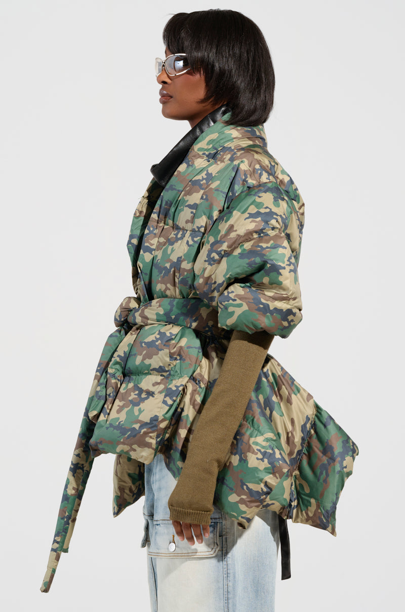 A person wearing the CAMO LONG RIB SLEEVE PEPLUM PUFFER COAT with a belt stands in profile. They have short black hair and wear glasses. The coat's olive green ribbed sleeves extend past their fingertips. They complete their outfit with light blue jeans against a plain background.
