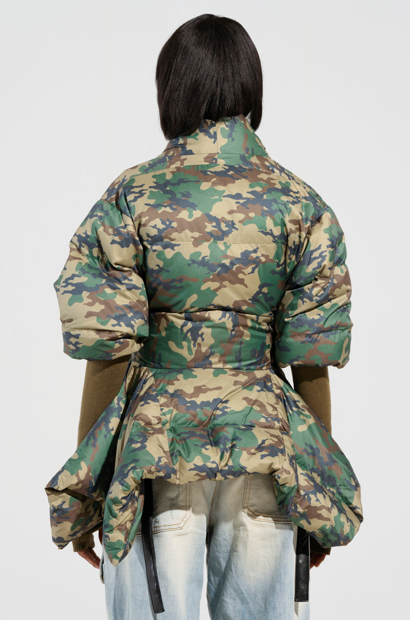 A person with dark hair, facing away, is dressed in the CAMO LONG RIB SLEEVE PEPLUM PUFFER COAT, showcasing exaggerated shoulders and a peplum hem, paired with light denim jeans against a plain white background.