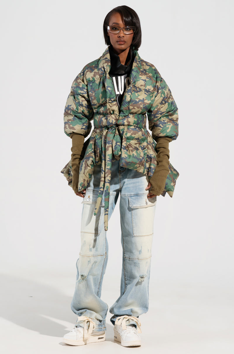 A person stands confidently in a fashionable CAMO LONG RIB SLEEVE PEPLUM PUFFER COAT, featuring ribbed sleeves and a belt. They're styled with distressed light blue jeans, white sneakers, and a black shirt underneath. They complete the look with glasses and maintain a neutral expression.