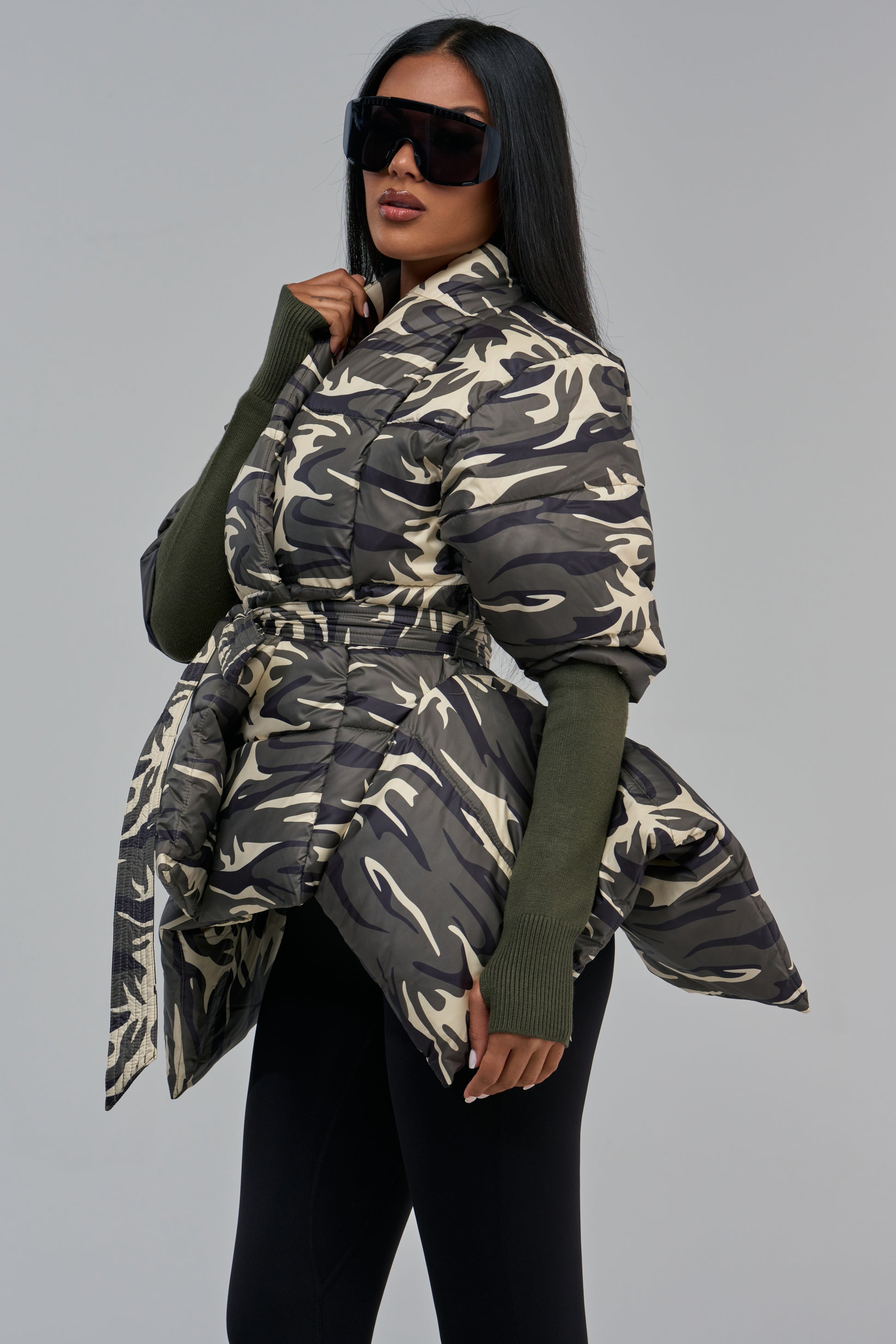 A person wears a RIB SLEEVE PEPLUM PUFFER IN CAMO, a camouflage jacket designed with long ribbed sleeves and a belted waist.