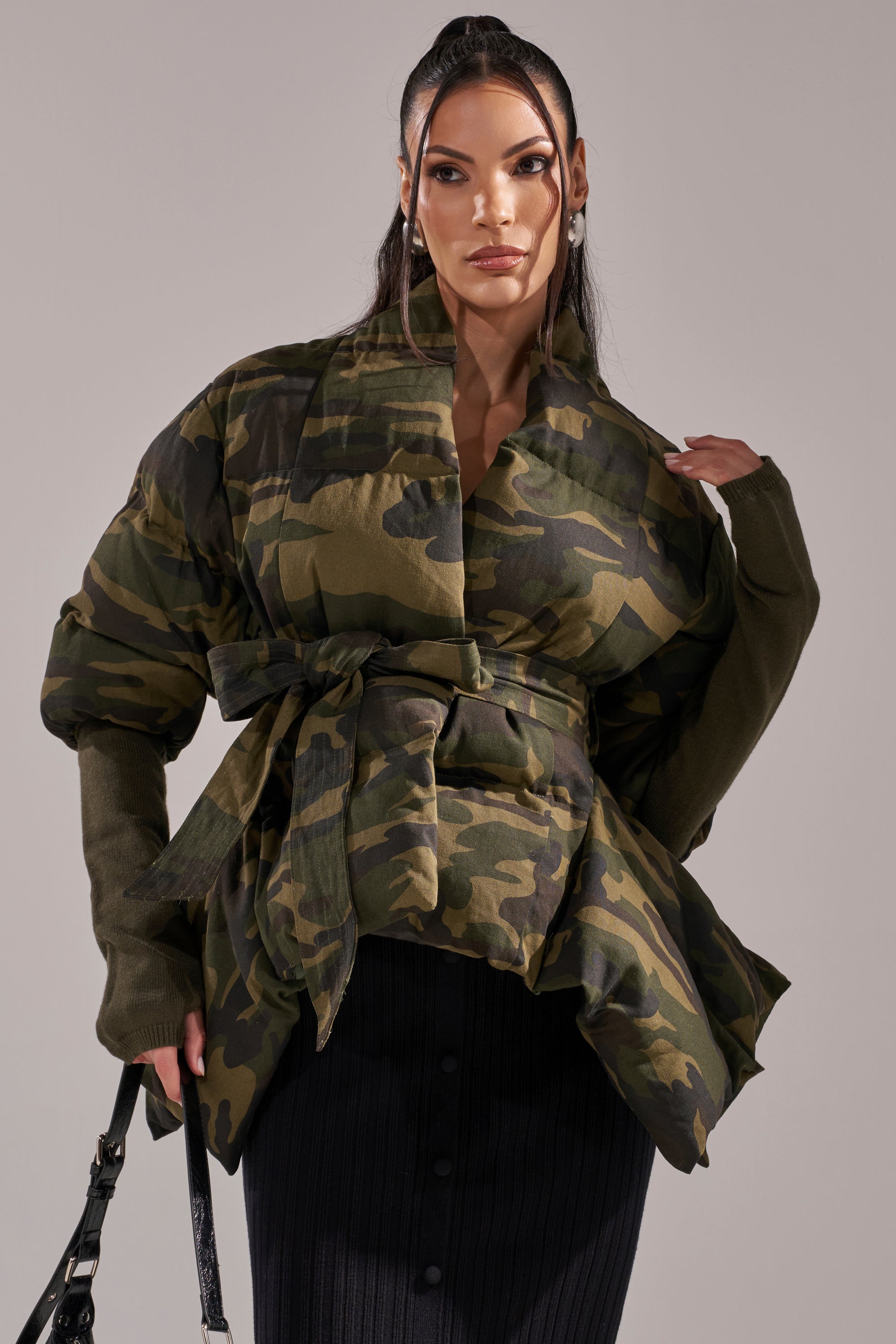 A woman models the RIB SLEEVE PEPLUM PUFFER IN CAMO, an oversized belted jacket with wide, ribbed sleeves, styled over a black buttoned skirt. Her long dark hair is pulled back and she accessorizes with large earrings and a black strap in hand. 0