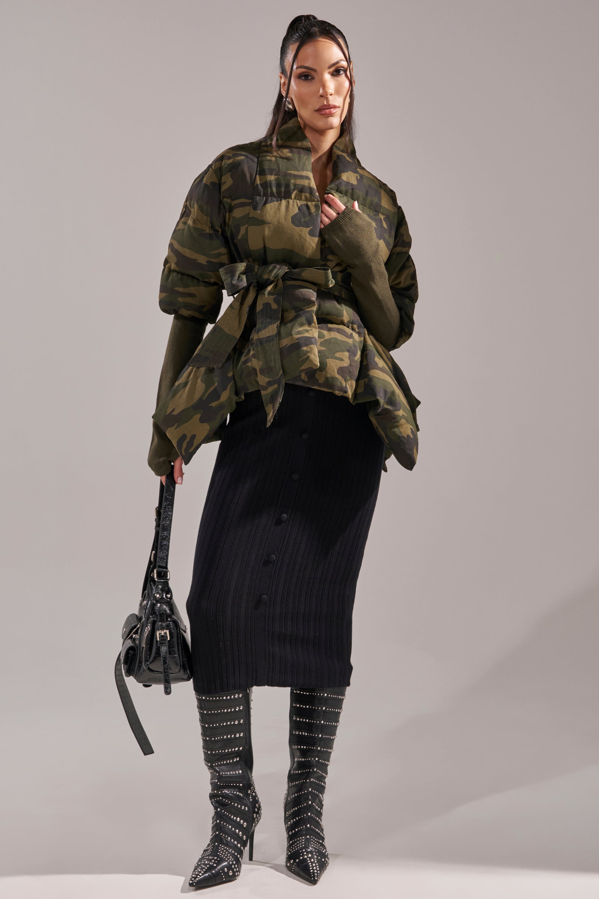 A woman stands confidently in the RIB SLEEVE PEPLUM PUFFER IN CAMO jacket, paired with a black ribbed midi skirt with buttons, studded knee-high boots, and a black handbag against a plain gray background. 9