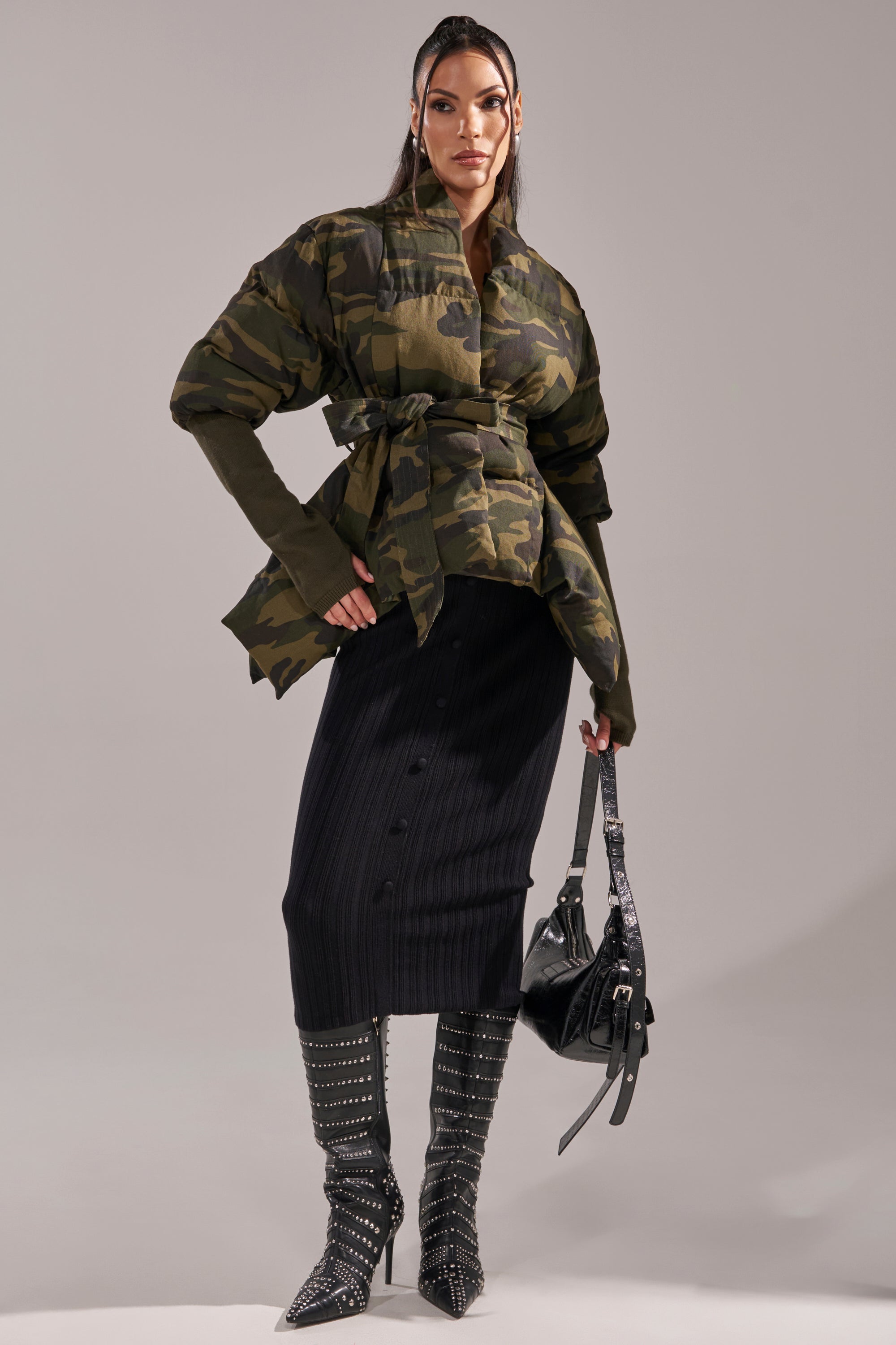 A woman poses in the RIB SLEEVE PEPLUM PUFFER IN CAMO, paired with a long black ribbed skirt and studded knee-high boots. She carries a black handbag, her hair straight and sleek, against a neutral background. 10