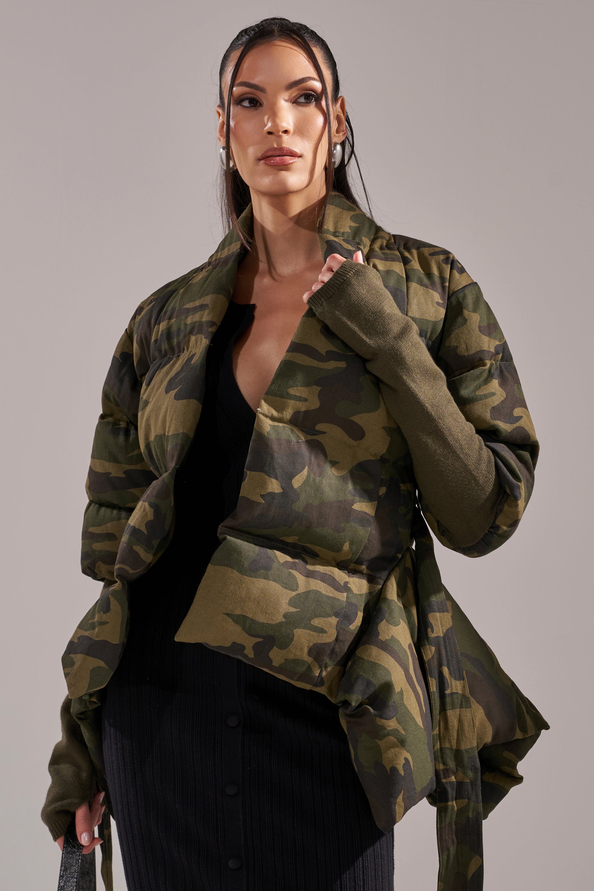 A woman wears the RIB SLEEVE PEPLUM PUFFER IN CAMO over a black top and skirt, standing against a neutral background with straight hair framing her face as she looks confidently ahead. 1