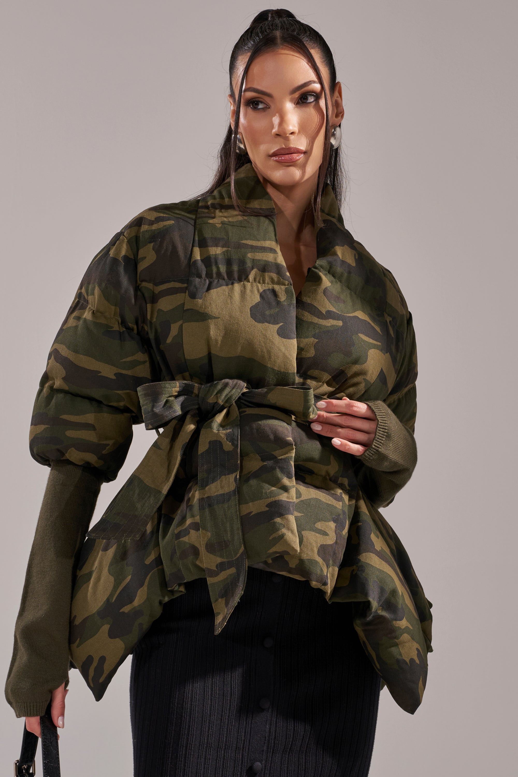 A woman poses confidently in the RIB SLEEVE PEPLUM PUFFER IN CAMO, featuring a green camouflage design, peplum silhouette with a waist tie, and olive rib sleeves, styled with a dark buttoned ribbed skirt. 2