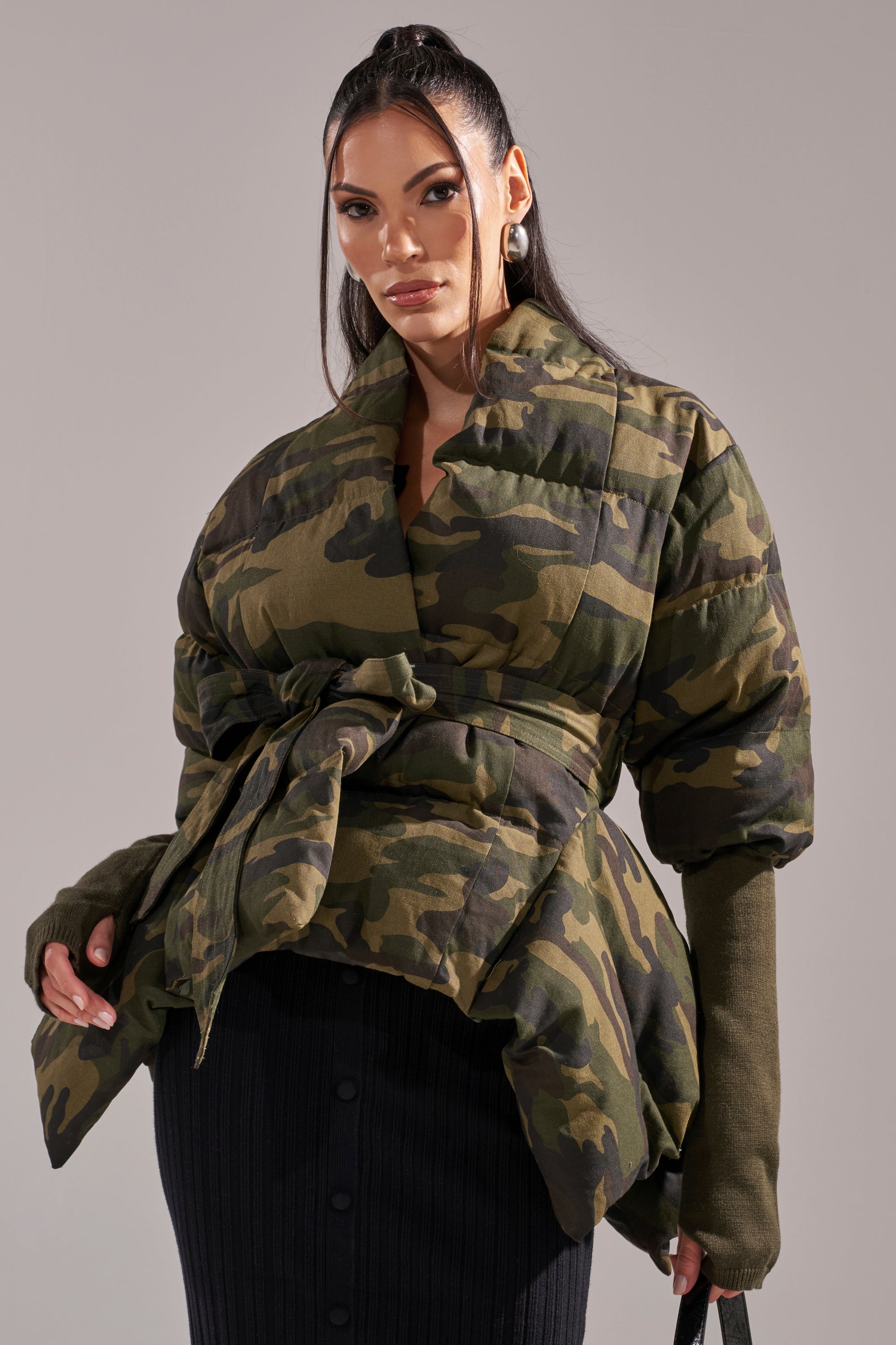 A woman wears the RIB SLEEVE PEPLUM PUFFER IN CAMO over a black buttoned skirt, posing against a plain light gray background. 3