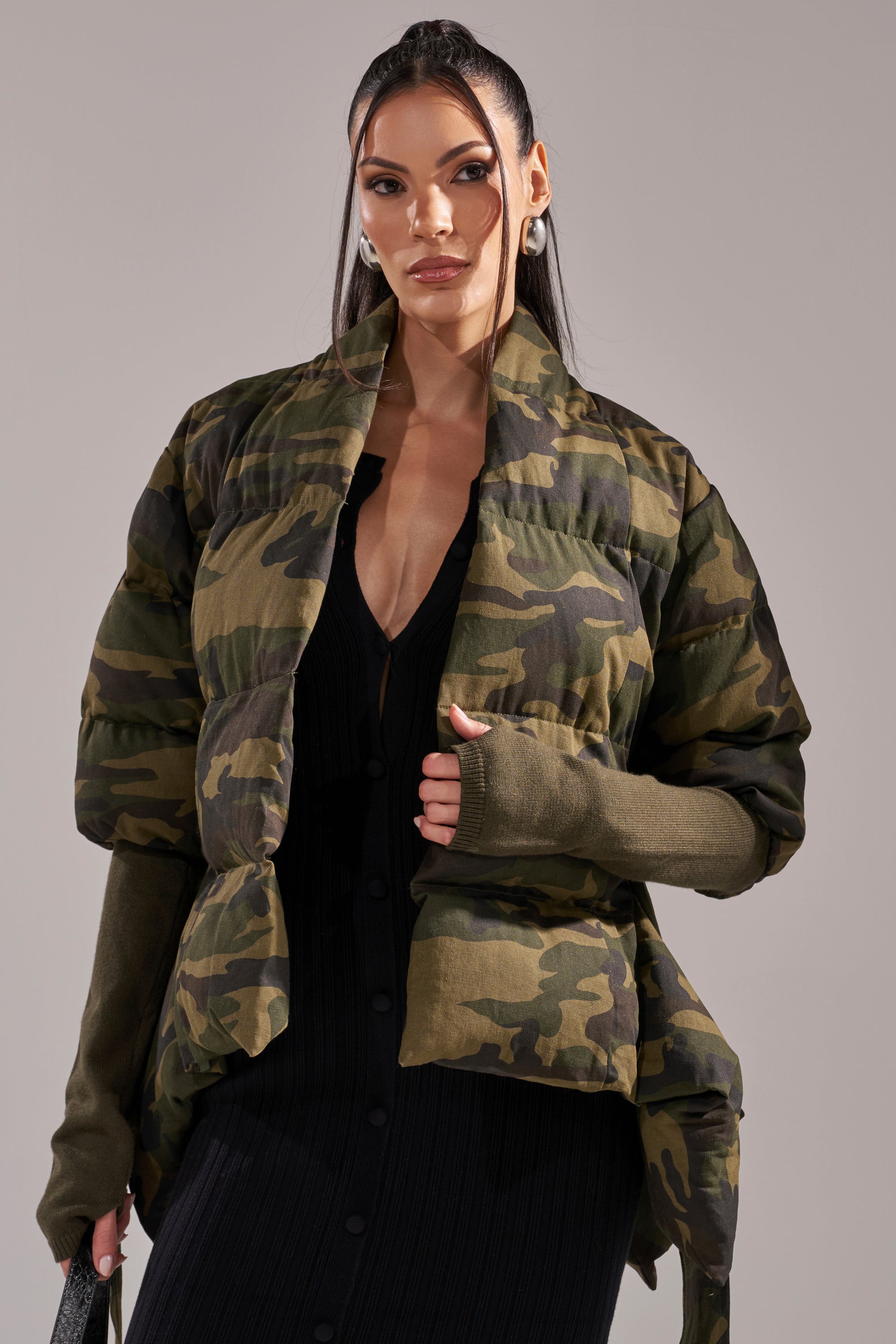 A woman with long dark hair wears the RIB SLEEVE PEPLUM PUFFER IN CAMO over a black button-front dress, standing confidently against a plain light background and looking directly at the camera. 4