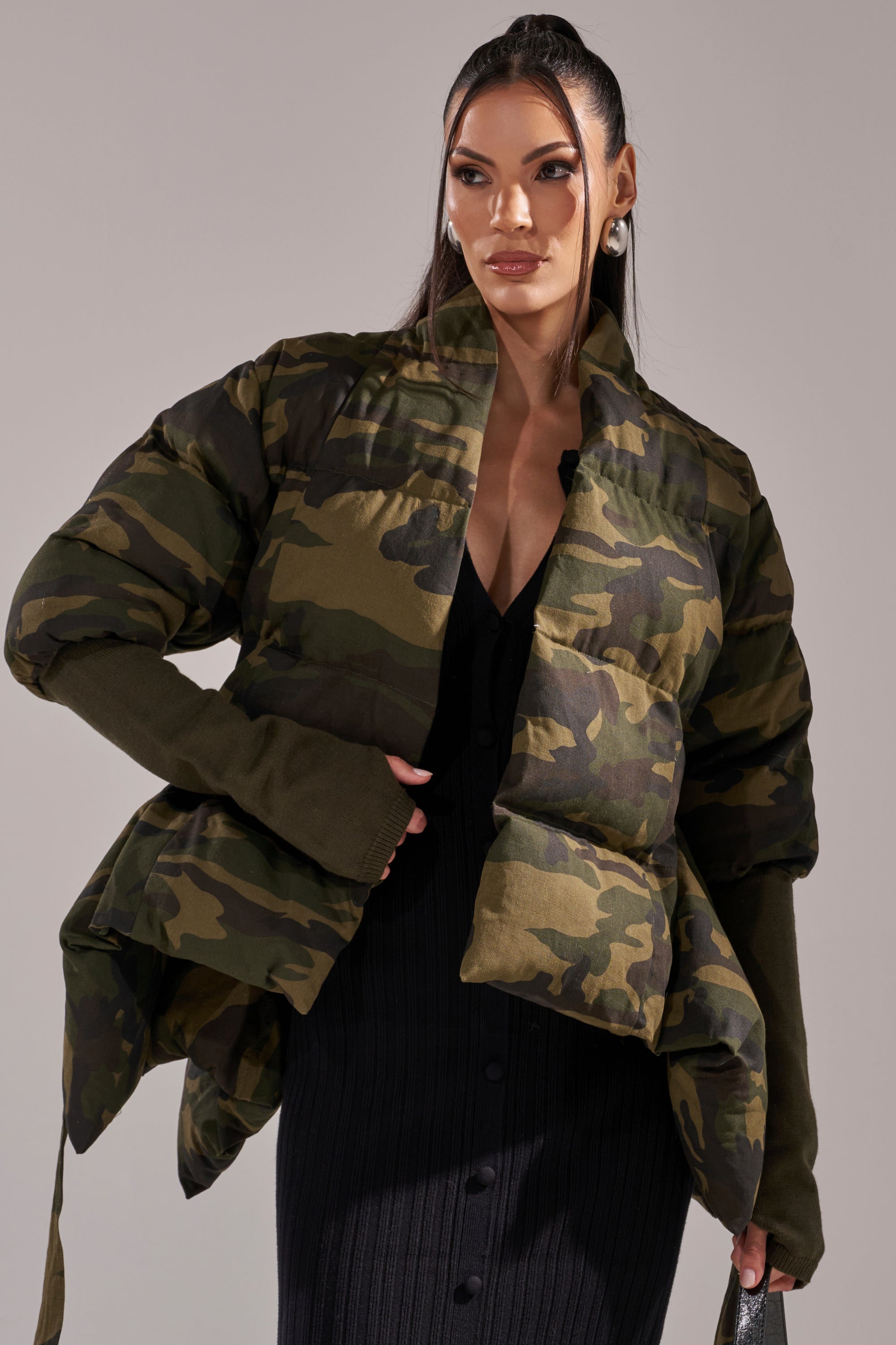 A woman stands confidently, wearing the RIB SLEEVE PEPLUM PUFFER IN CAMO over a black ribbed dress with button details, holding a bag and looking to the side. Her long hair is styled in a sleek ponytail. 5