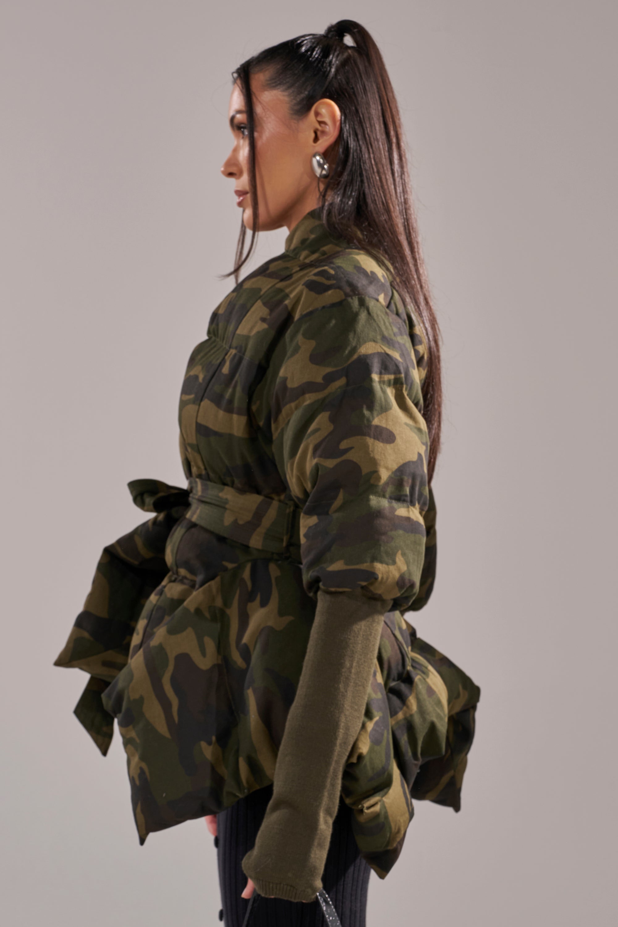 A woman with long, straight hair in a high ponytail models the RIB SLEEVE PEPLUM PUFFER IN CAMO, featuring voluminous rib sleeves and a belted waist, paired with dark gloves and posed in profile against a plain backdrop. 6