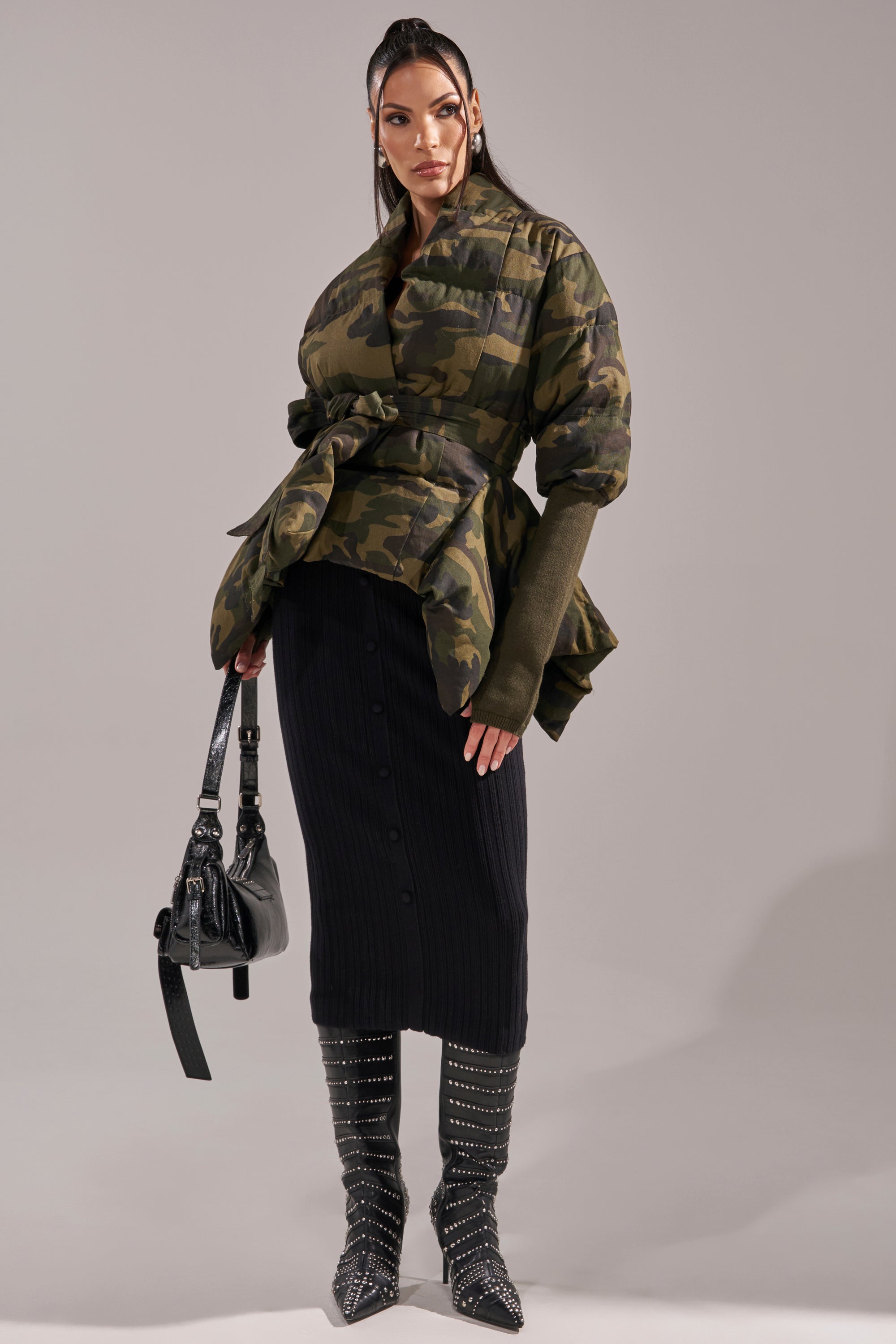 A woman poses in the RIB SLEEVE PEPLUM PUFFER IN CAMO, styled with a long black buttoned skirt, studded knee-high boots, and a black handbag against a plain gray backdrop. 8
