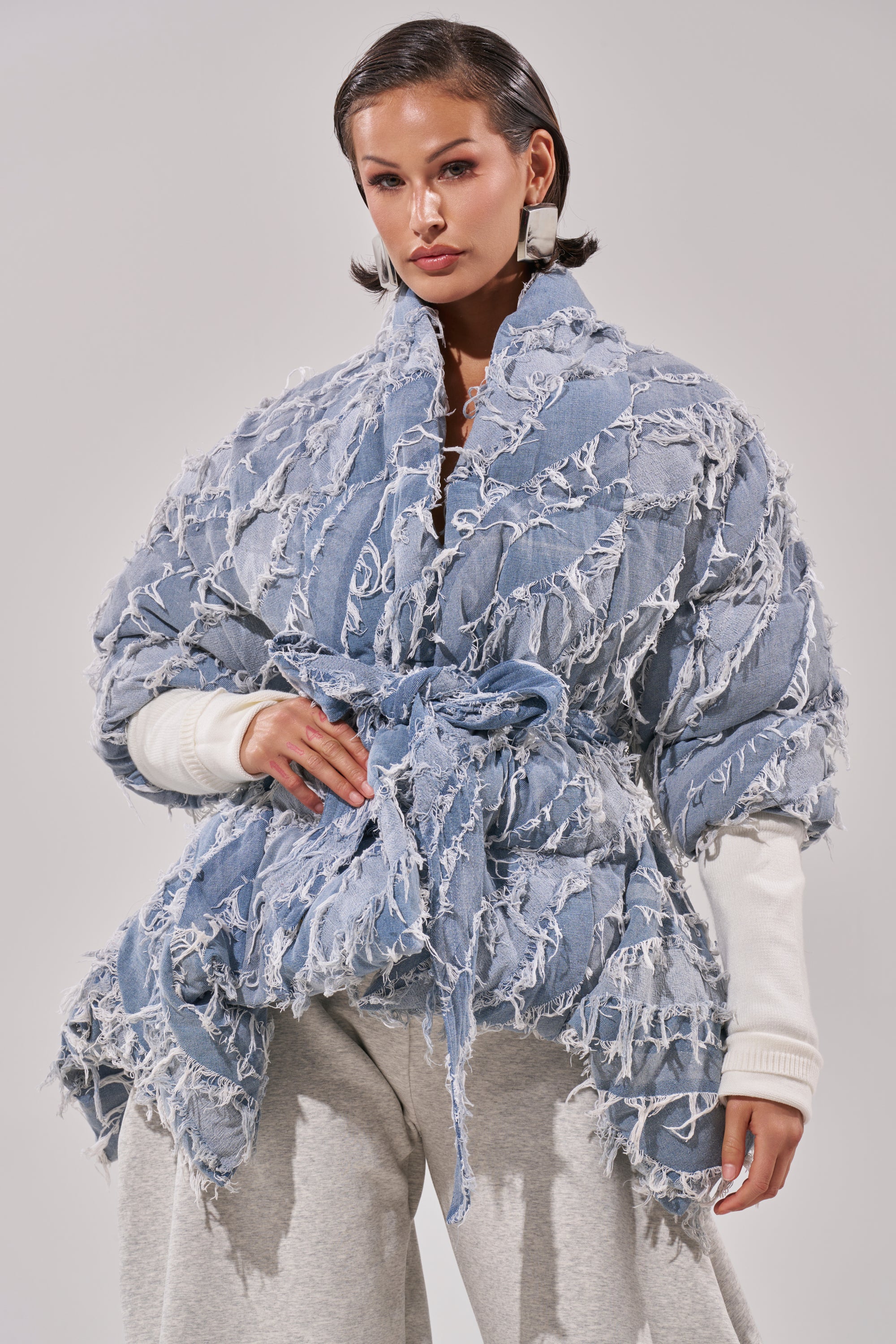 A woman styles the RIB SLEEVE PEPLUM PUFFER IN DENIM over a white top and light gray pants. Her short, sleek hair and statement earrings add a modern twist to this textured, fashion-forward outfit.