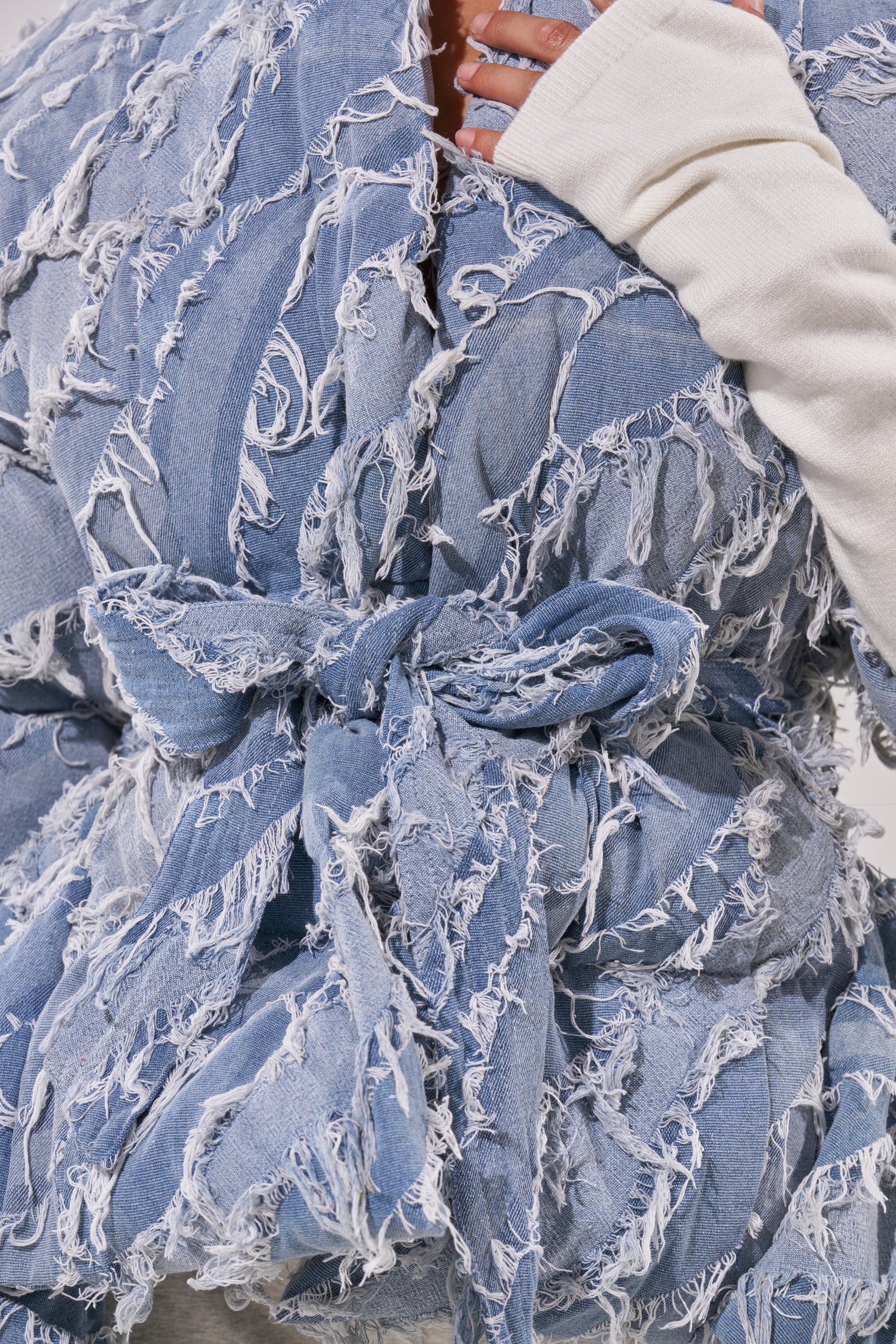 A close-up of a person wearing the RIB SLEEVE PEPLUM PUFFER IN DENIM, featuring textured, fringed patchwork, tied at the waist with a matching belt over a white long-sleeve top for an elevated street-luxe style.