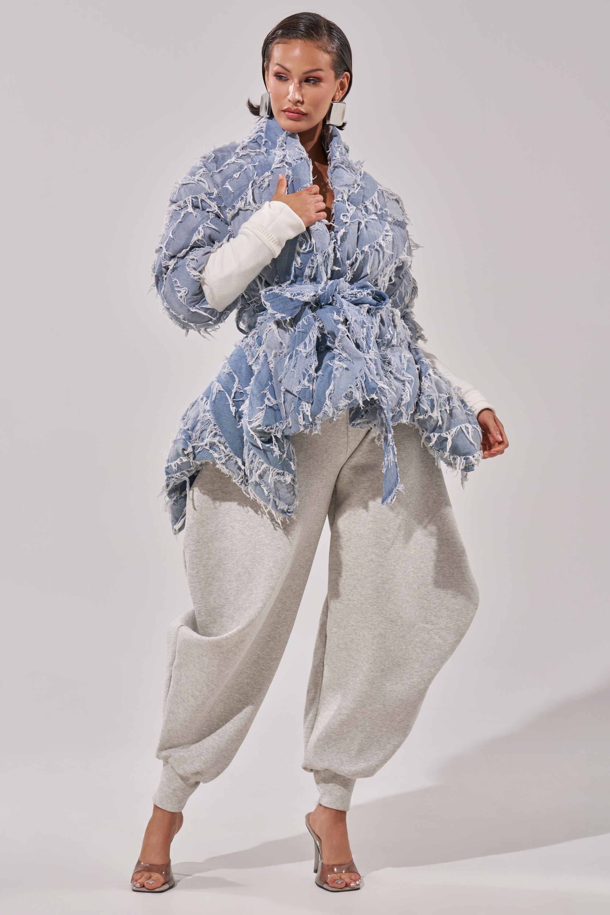 A woman poses in the RIB SLEEVE PEPLUM PUFFER IN DENIM, layered over a white top with loose light gray sweatpants and clear heeled sandals, set against a plain, light background.