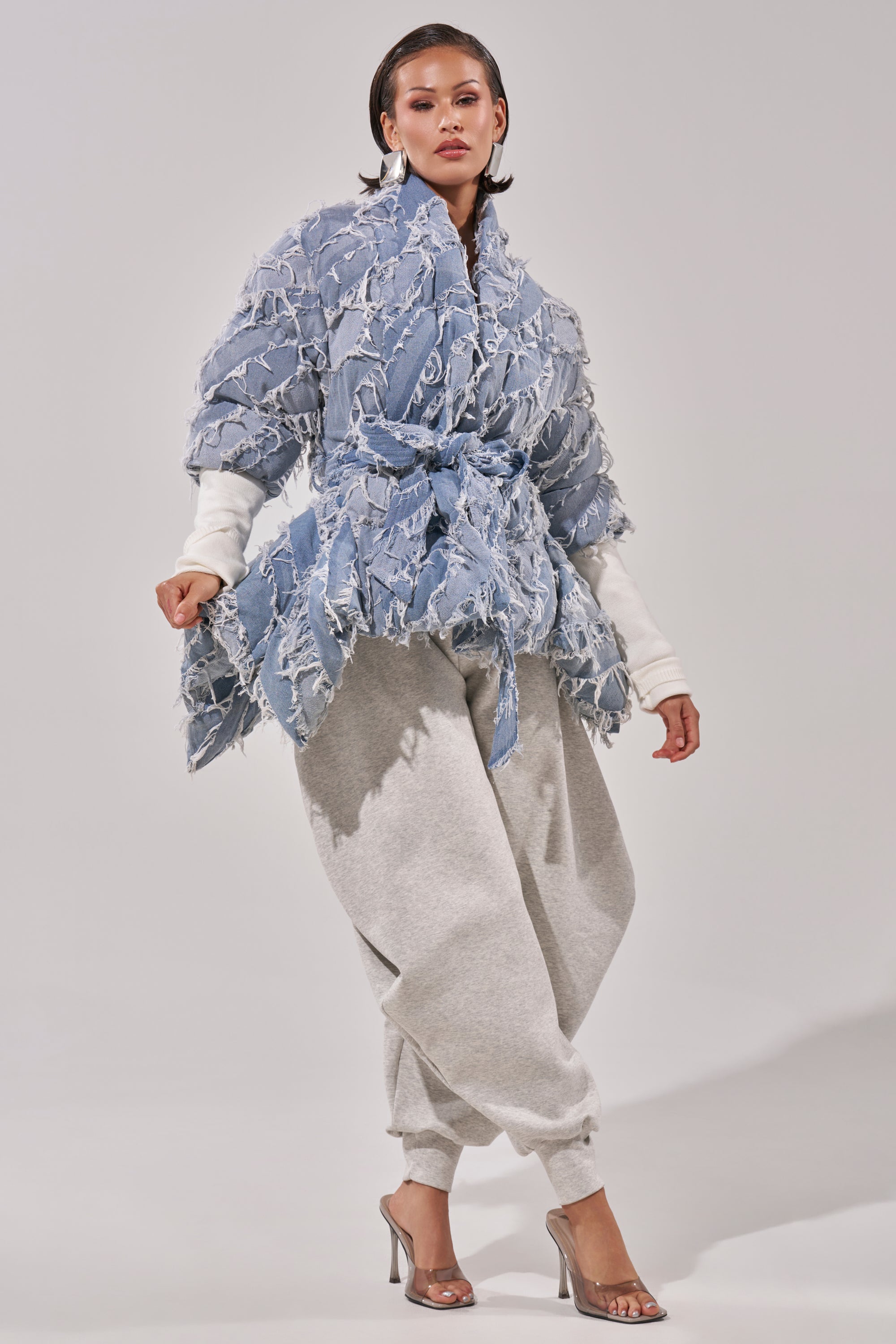 A woman poses in the RIB SLEEVE PEPLUM PUFFER IN DENIM tied at the waist over a white top, paired with loose gray sweatpants and transparent high-heeled sandals against a plain background.