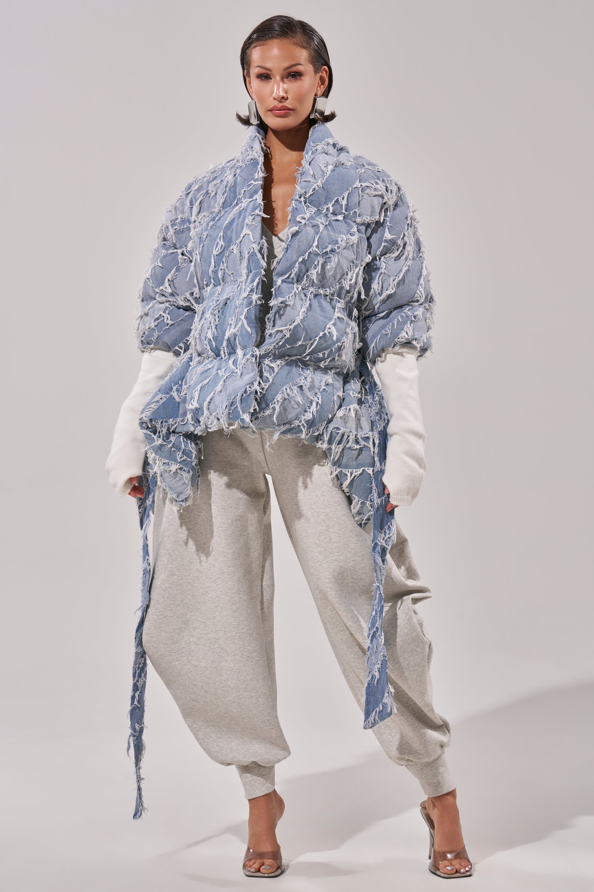 A woman models the RIB SLEEVE PEPLUM PUFFER IN DENIM, featuring textured light blue fabric and long ties, styled over white sleeves and loose gray sweatpants with clear high-heeled shoes against a plain backdrop.