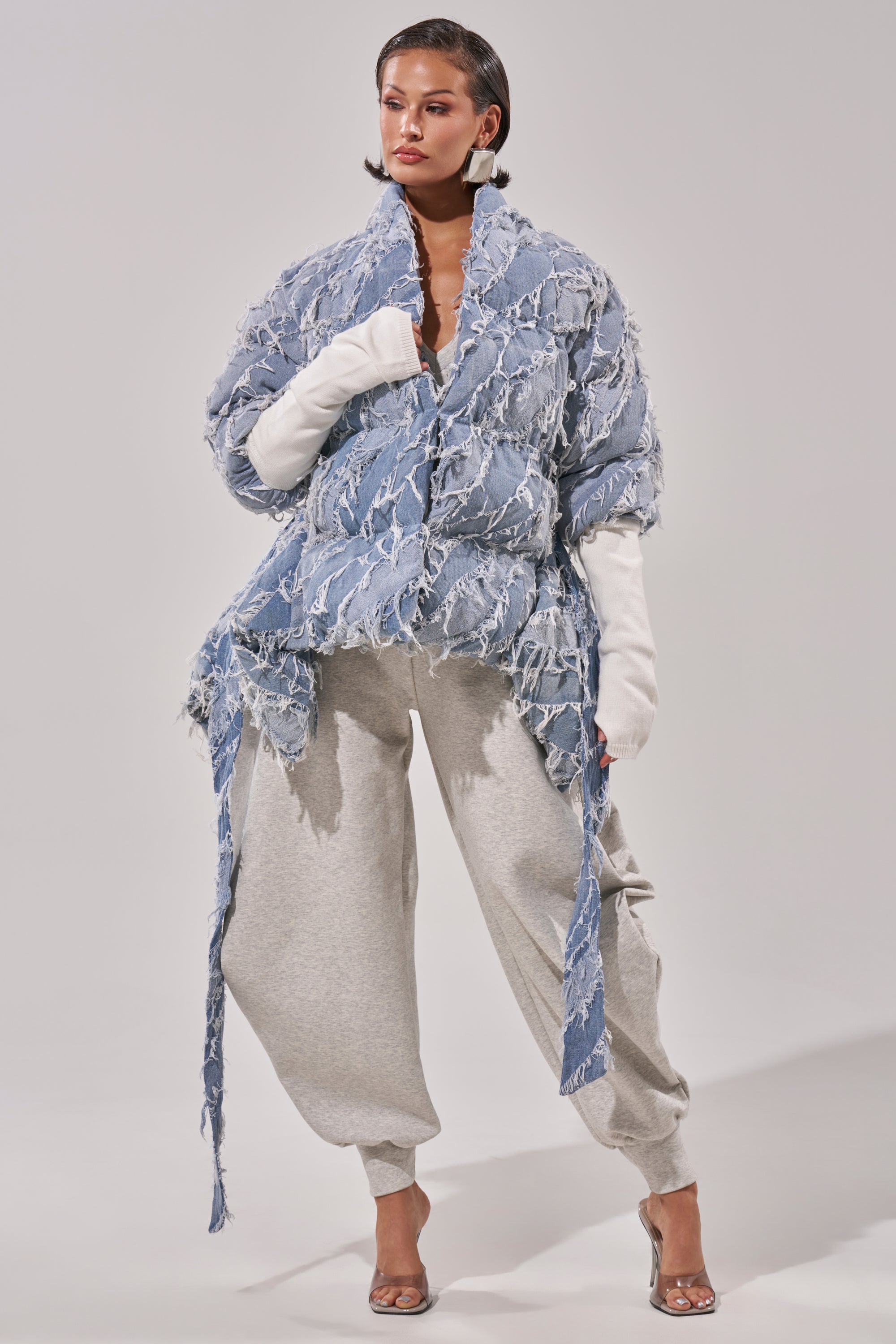 A woman models the RIB SLEEVE PEPLUM PUFFER IN DENIM, styled with oversized white gloves, gray sweatpants, and clear high-heeled sandals against a plain background.