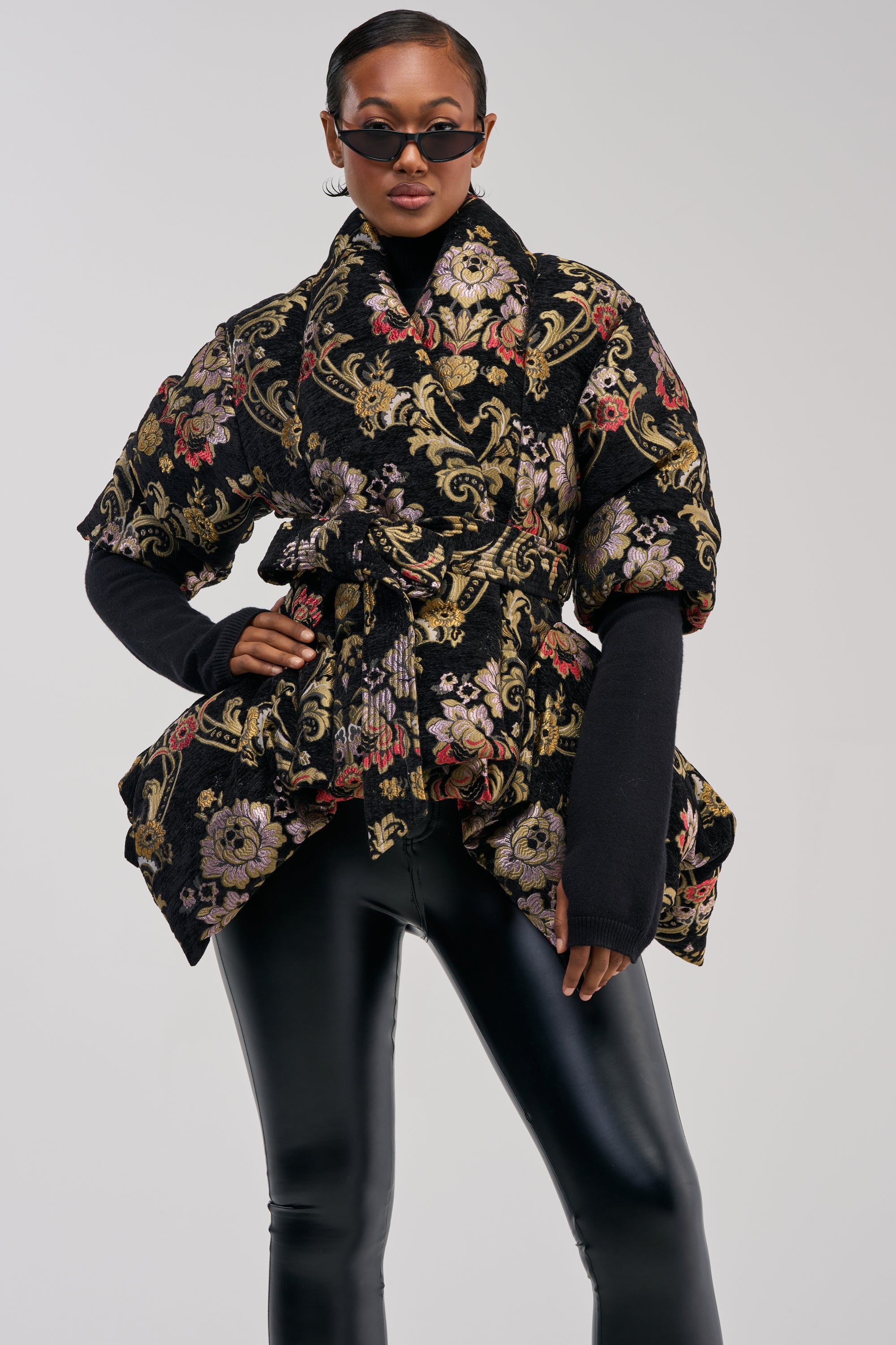 A woman poses confidently against a plain background, dressed in dark sunglasses and sleek black leather pants, while her boldly sophisticated look is elevated by the MONET JACQUARD PEPLUM PUFFER—a statement jacket with an artful jacquard texture and a flattering peplum silhouette.