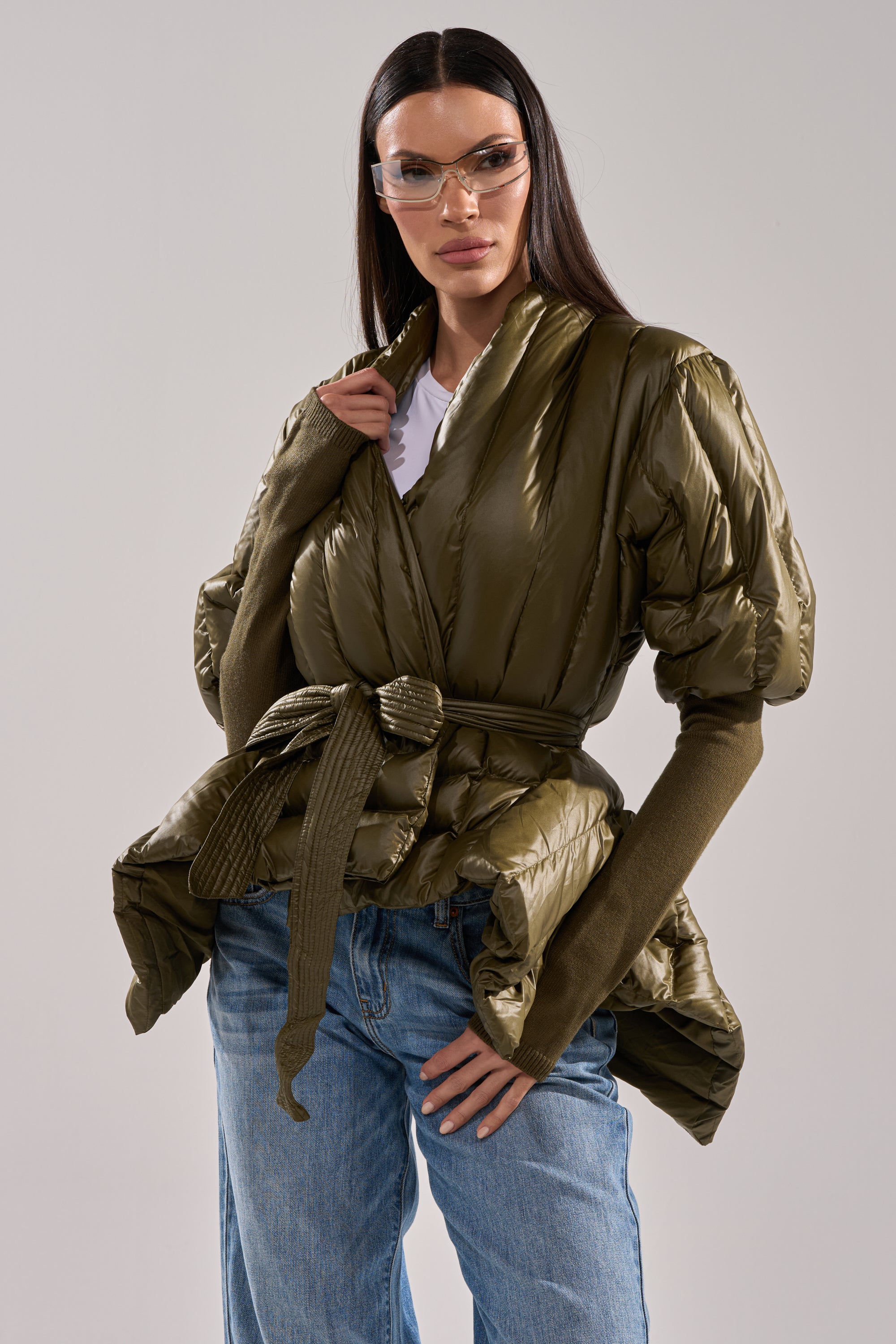 A woman wearing clear glasses, the RIB SLEEVE PEPLUM PUFFER IN OLIVE with a tie belt, a brown long-sleeve top, and blue jeans stands confidently against a plain light gray background. 1
