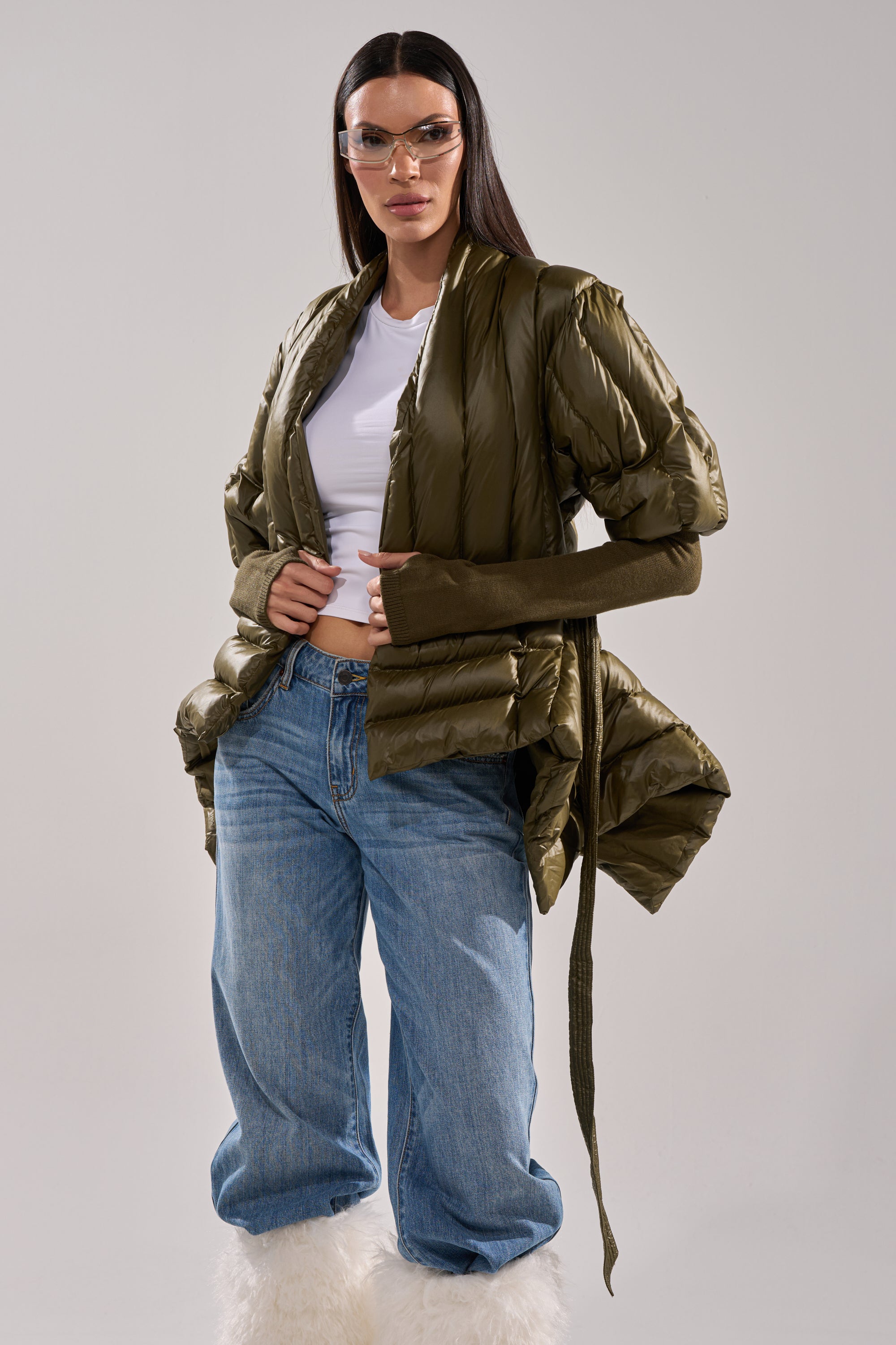 A woman models the RIB SLEEVE PEPLUM PUFFER IN OLIVE with a white crop top, blue jeans, long brown sleeves, oversized clear glasses, and white furry boots against a plain background. 2
