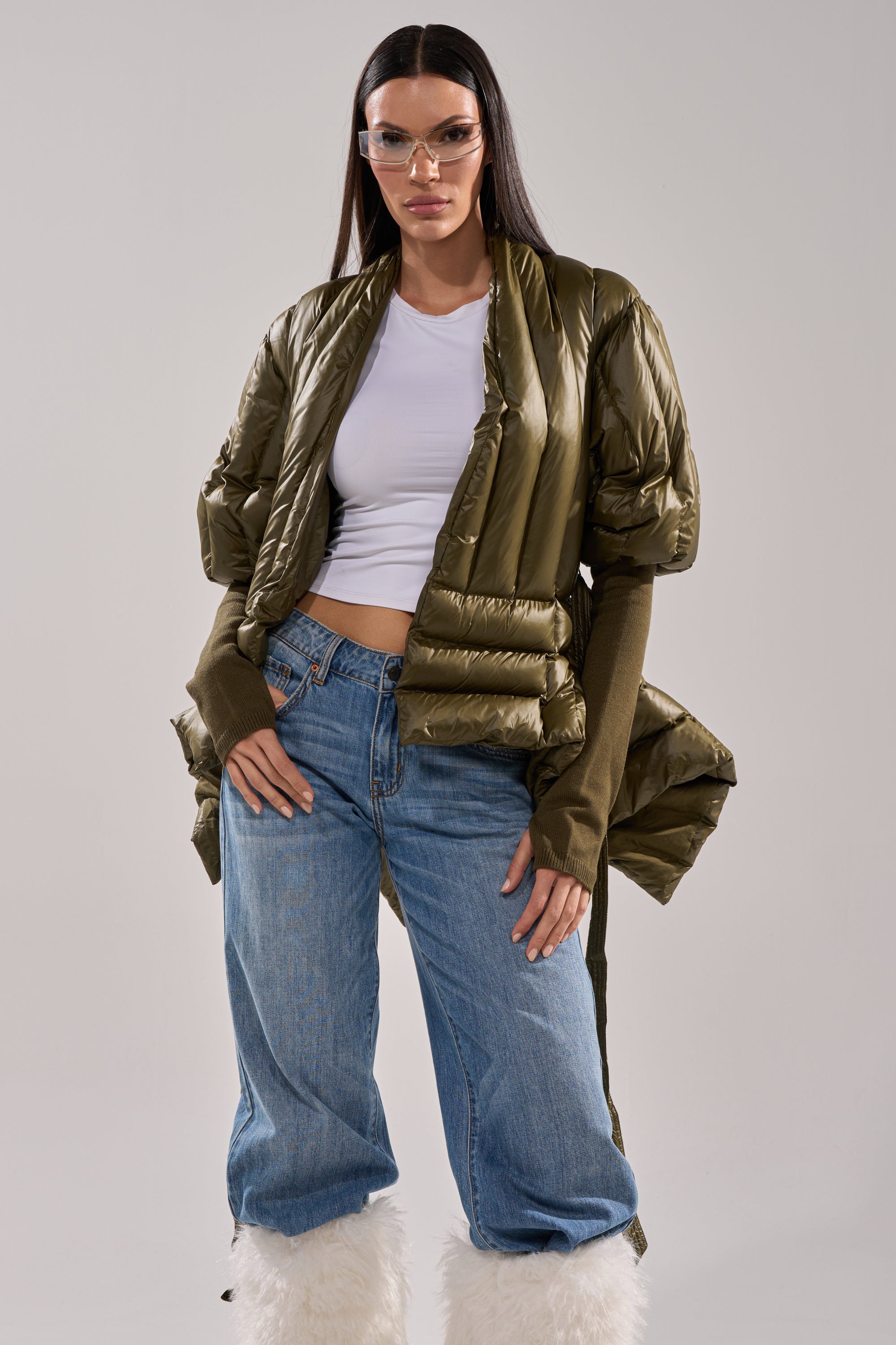 A woman poses confidently against a plain background in the RIB SLEEVE PEPLUM PUFFER IN OLIVE, styled with a white crop top, blue jeans with white furry cuffs, long brown fingerless gloves, and clear visor glasses. 3