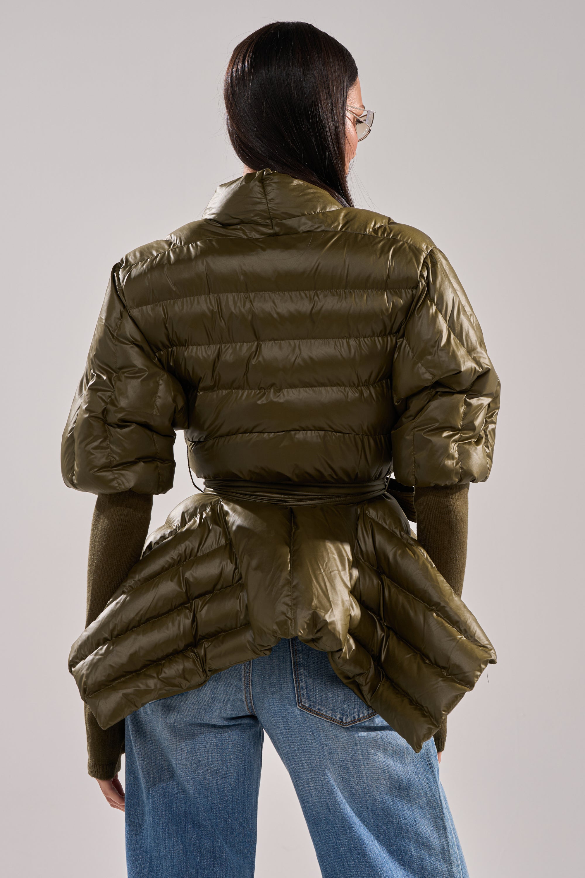 A person with long dark hair and glasses, seen from behind against a neutral background, wears blue jeans and the RIB SLEEVE PEPLUM PUFFER IN OLIVE, which features an exaggerated silhouette. 5