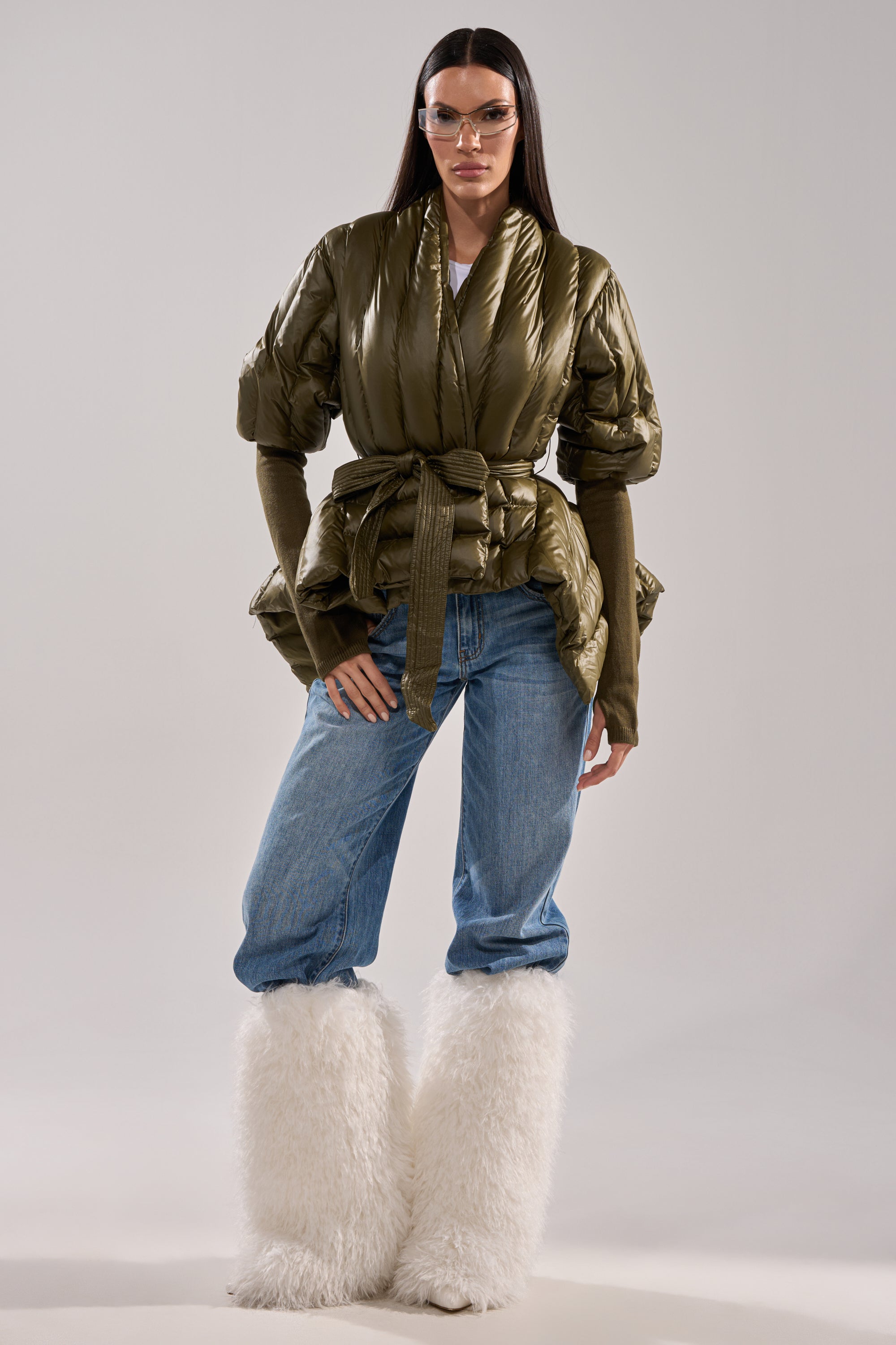 A woman stands against a plain background, wearing clear goggles, blue jeans, fluffy white boots, and the RIB SLEEVE PEPLUM PUFFER IN OLIVE. 6