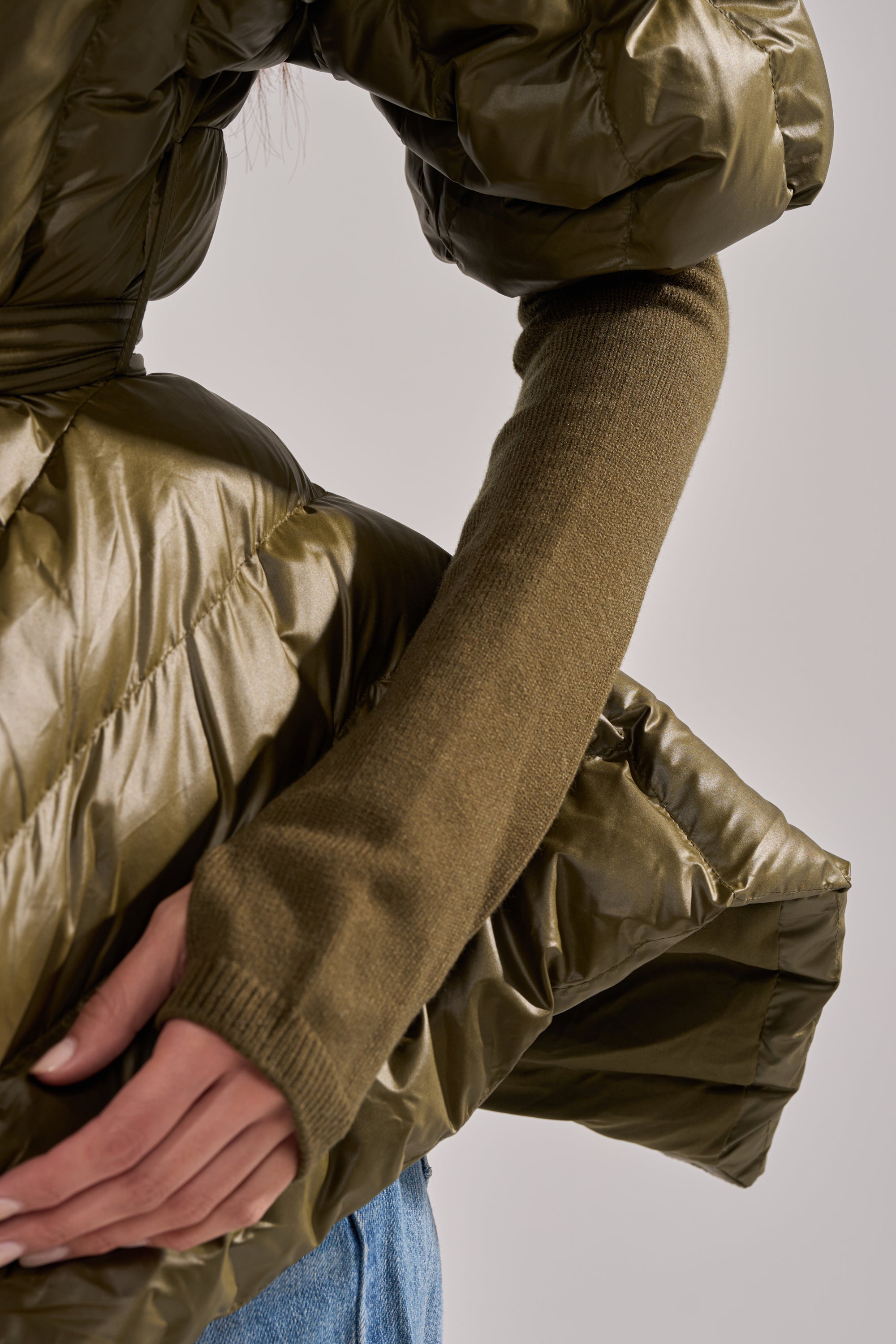 A close-up shows a person in the RIB SLEEVE PEPLUM PUFFER IN OLIVE, featuring ribbed sleeves and a peplum design. One hand rests on their hip, styled with blue jeans against a plain, light background. 8