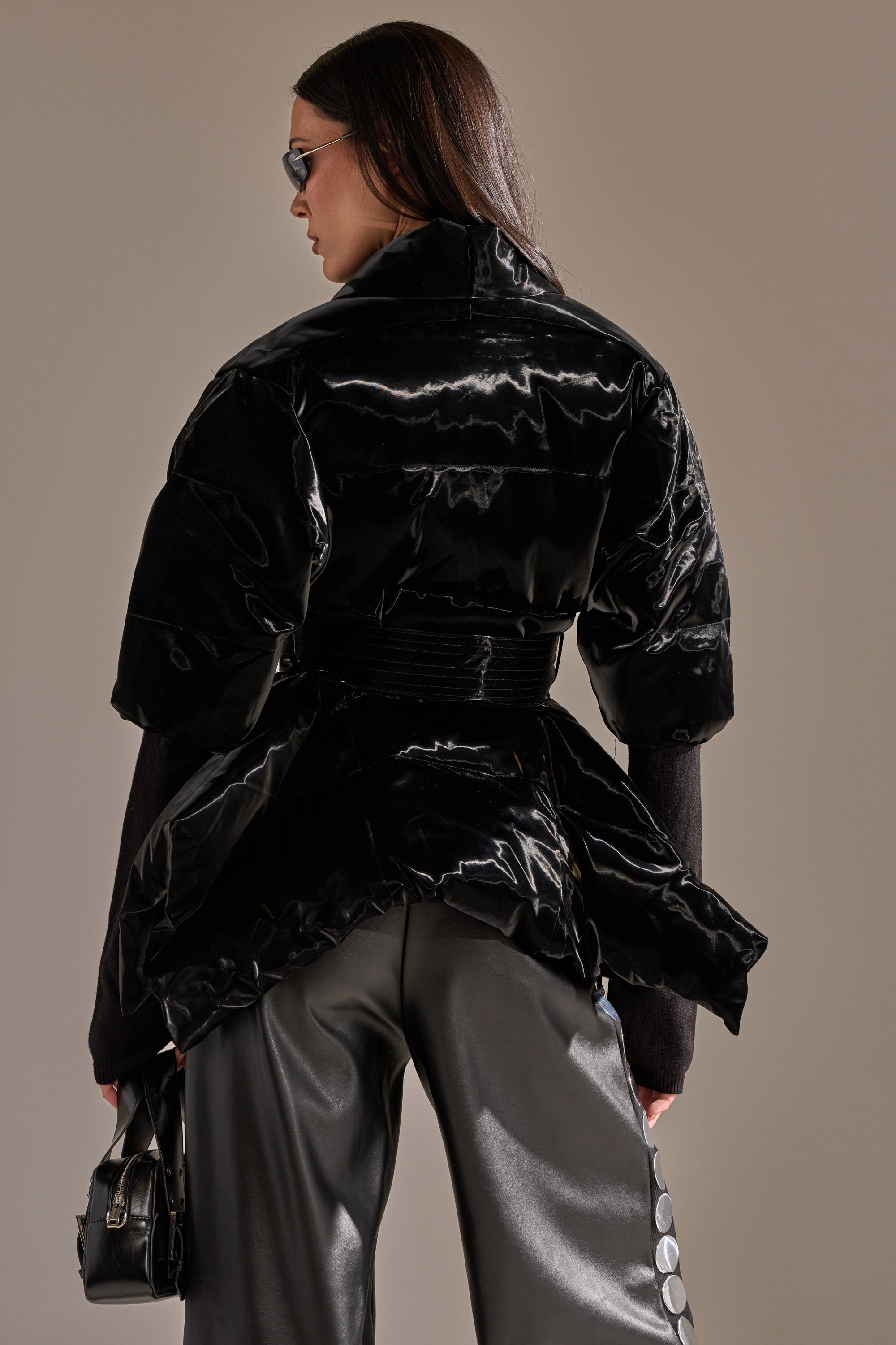 A person stands facing away in the RIB SLEEVE PEPLUM PUFFER IN LIQUID VELVET, paired with black leather pants and sunglasses, holding a small handbag against a plain neutral background. 4