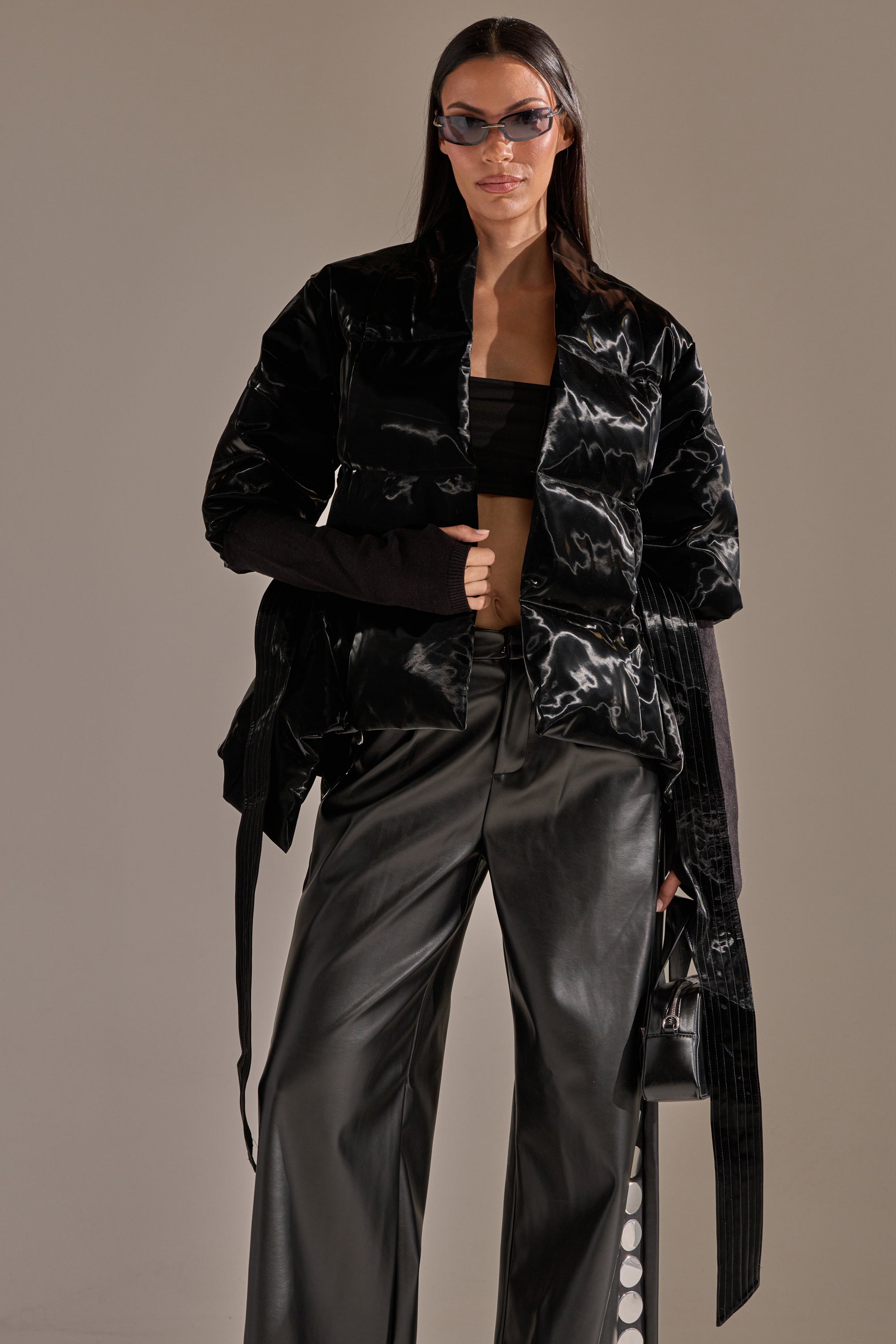 A woman confidently poses in the RIB SLEEVE PEPLUM PUFFER IN LIQUID VELVET, wide-leg black leather pants, a black crop top, angular sunglasses, and holds a small black handbag. 5