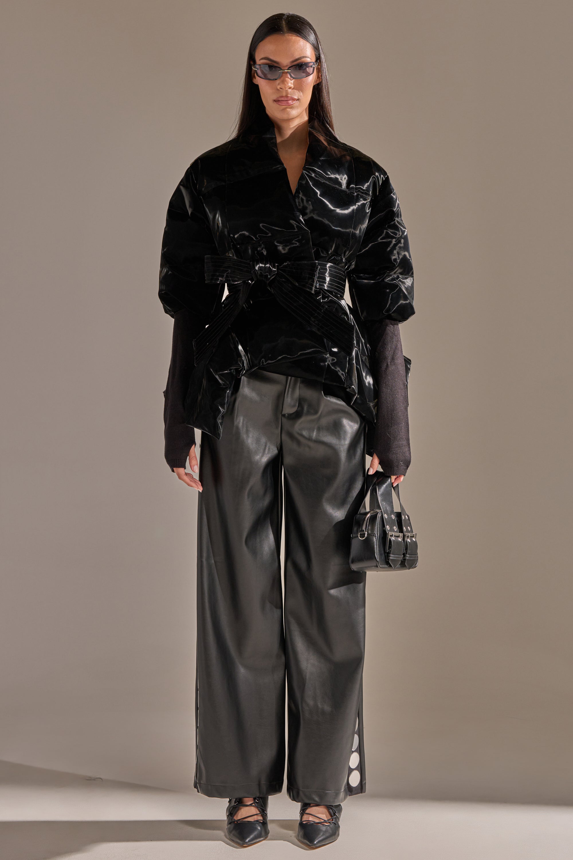 A person stands facing forward in the RIB SLEEVE PEPLUM PUFFER IN LIQUID VELVET, paired with loose black leather pants, black shoes, sunglasses, and a small black handbag. The background is plain and neutral. 6